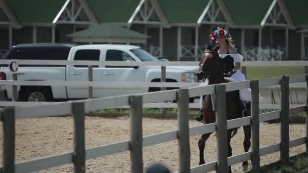 Minsk, Bielorrússia - 19 de julho de 2019: Duas jovens garotas de vestido vintage medieval branco com um espartilho e com grinaldas em suas cabeças andam juntas a cavalo em um rancho — Vídeo de Stock