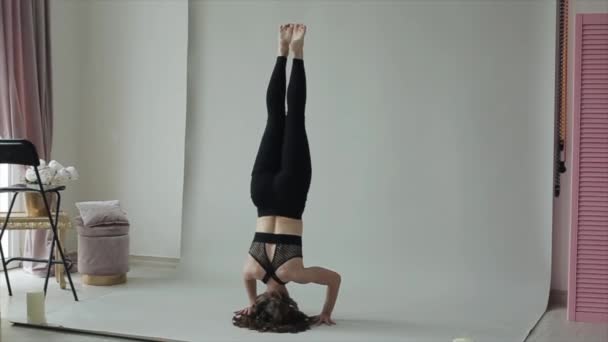 Young sports girl practices yoga poses, standing on her head and lowering her legs to the floor in a black top and leggings — Stock Video