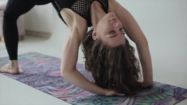 Hermosa joven practica yoga en casa y toma la pose de un puente mientras está de pie sobre una alfombra en ropa deportiva negra. Primer plano — Vídeos de Stock