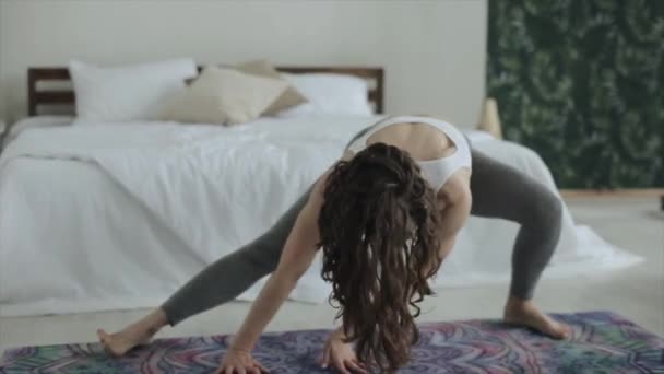 Joven chica atractiva practica yoga mientras hace diferentes ejercicios en la estera en su habitación en ropa deportiva hermosa — Vídeos de Stock