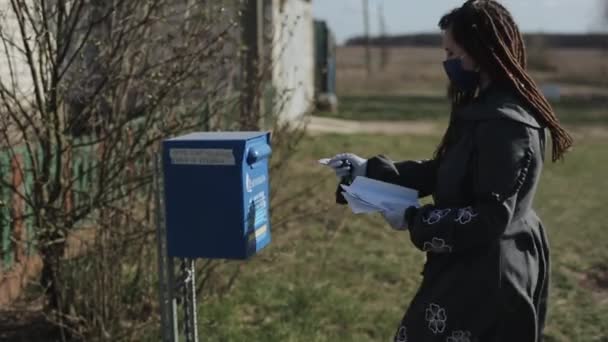 ベラルーシのBobrisk - 2020年4月20日:ロシアの村の通りの郵便ポストに少女が手紙を投げます。パンデミックの間、ドレッドロックと保護マスクと手袋を持つ少女。サイドビュー — ストック動画