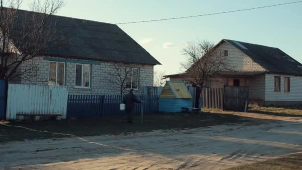 Bobruisk, Bielorrusia - 20 de abril de 2020: Un anciano con un cubo va al pozo a lo largo de una calle del pueblo, apoyado en un bastón. Pueblo ruso — Vídeos de Stock