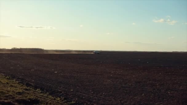 Egy autó halad végig egy vidéki úton szántott mezők között a kék ég felé, és egy porfelhőt vet fel a kerekek alól. Oldalnézet