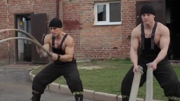 Two young men train and do different exercises with battle ropes while standing outdoors. Crossfit. Close-up. The camera moves up and down — Stock Video