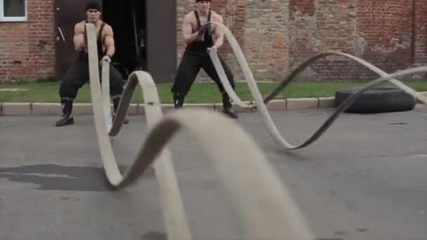 Dva mladí svalnatí muži cvičí a cvičí křížové cvičení s bojovými lany. Kamera se pohybuje nahoru a dolů za vlnami lan — Stock video