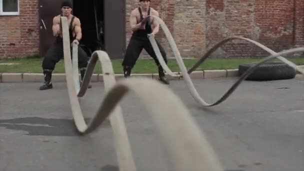 Twee jonge gespierde mannen trainen en doen crossfit oefeningen met gevechtstouwen. De camera beweegt op en neer achter de golven van touwen — Stockvideo