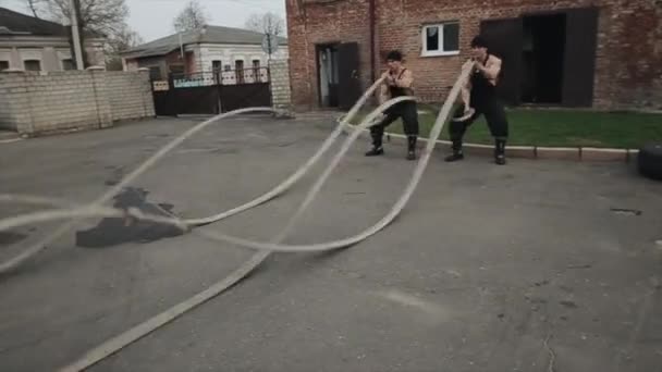 Two young muscular men doing crossfit training with battle ropes at high speed. The camera monitors the movement of men — Stock Video