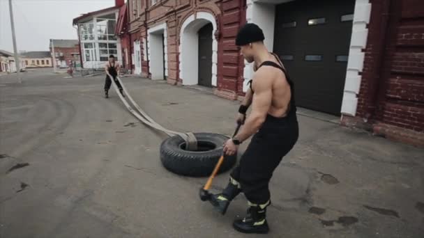 Två muskulösa unga män tränar och gör crossfitövningar med rep och hammare. Bakåt. Kameran följer männens rörelser — Stockvideo