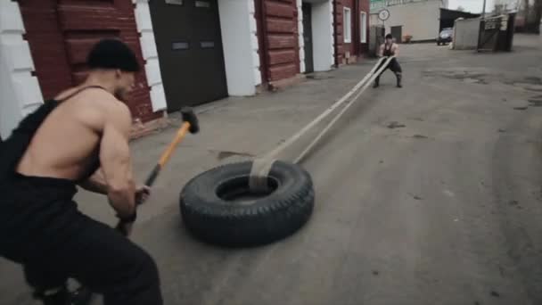 Dos jóvenes musculosos están entrenando y haciendo ejercicios de crossfit con un neumático y un martillo. Primer plano. La cámara sigue los movimientos de los hombres — Vídeo de stock