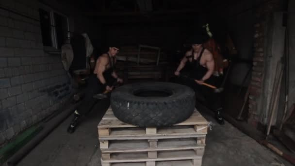 두 명 의근 육있는 청년 이 큰 망치 로 타이어 를 때리면서 훈련시키고 십 자형 운동을 한다. 클로즈업. 카메라는 앞뒤로 움직입니다. — 비디오