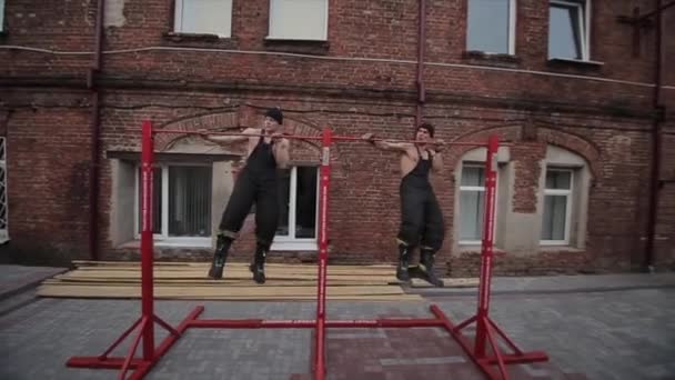 Twee jonge gespierde mannen trainen en doen extreme push-ups op de horizontale balk van links naar rechts. De camera beweegt van links naar rechts — Stockvideo