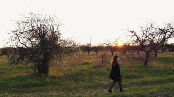 Вид с воздуха на молодую девушку, которая гуляет в весеннем саду под лучами заходящего солнца и весело танцует — стоковое видео