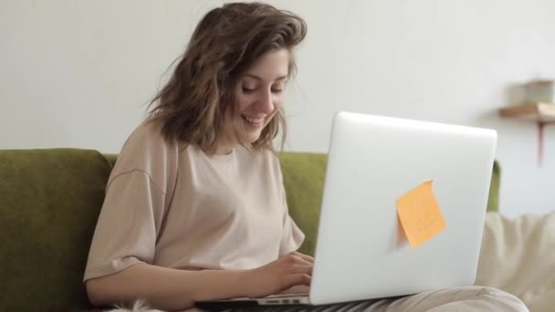 Fiatal gyönyörű lány szabadúszó dolgozik egy laptop, miközben otthon ül a kanapén mellett a kis kutya. Közelkép — Stock videók