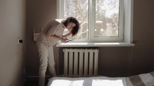 Menina bonita jovem corta as extremidades de seu cabelo com tesoura enquanto está perto da janela em seu quarto durante a quarentena Covid-19 — Vídeo de Stock