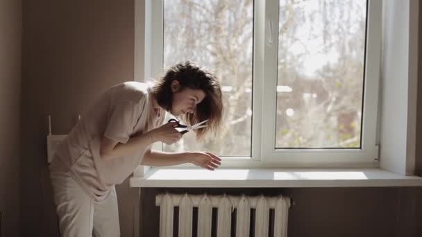 Una joven se para cerca de la ventana de su habitación y se corta las puntas del cabello con tijeras durante la cuarentena Covid-19 — Vídeos de Stock