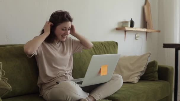 Giovane ragazza freelance che lavora a casa seduta sul divano e sorridente parlando con qualcuno sul computer portatile quando il suo cane salta a lei — Video Stock