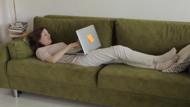 Молода дівчина працює вдома на ноутбуці під час карантину Covid 19 лежить на дивані у своїй кімнаті і розмовляє з кимось онлайн — стокове відео