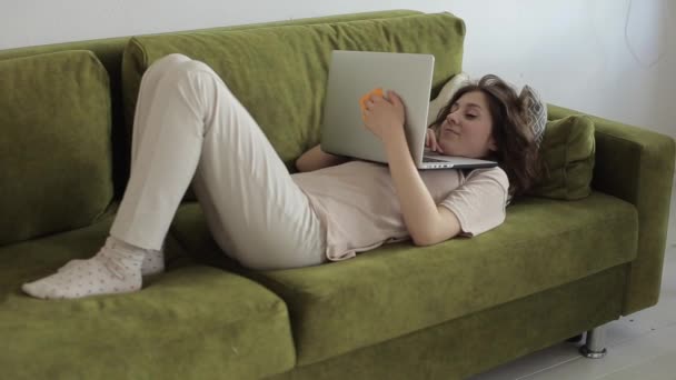 Uma jovem freelancer trabalha em casa em um laptop enquanto está deitada em um sofá em seu quarto durante a quarentena devido a uma pandemia de coronavírus. Close-up. engraçado detém computador — Vídeo de Stock