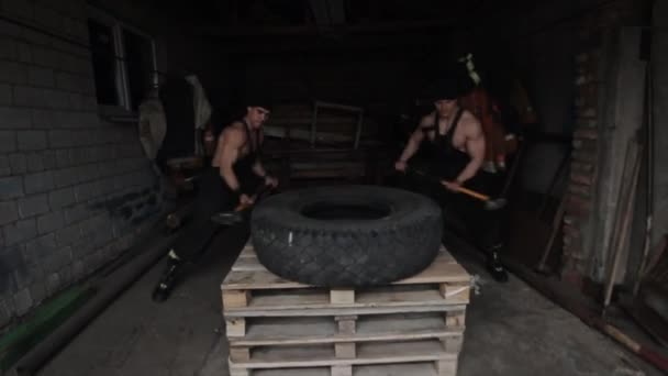 Dva svalnatí mladí muži trénují a dělají křížová cvičení, zatímco bijí pneumatiku kladivy. Kamera se pohybuje sem a tam. Detailní záběr — Stock video