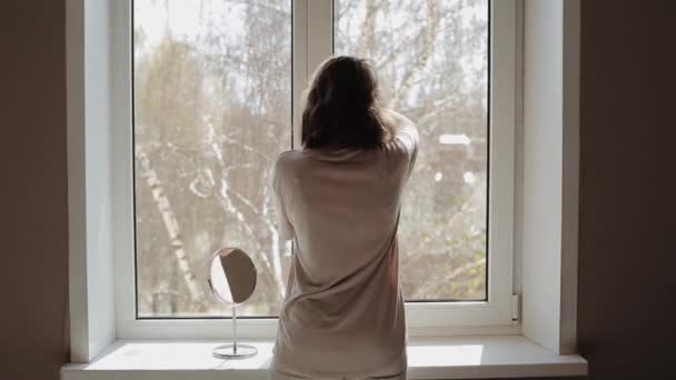 A young girl opens a window straightens her hair and enjoys a sunny day next to her little dog. Back view. Close-up — Stock Video