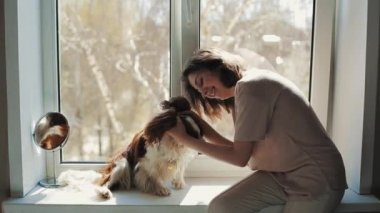 Pencere kenarında oturan genç bir kız köpeği Cavalier King Charles Spaniel 'e sarılıp öpüyor. Yakın plan.