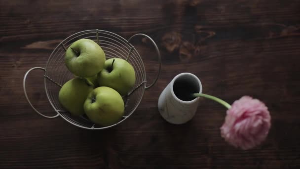 Вид зверху на кошик з зеленими яблуками і вазою з рожевою квіткою, що стоїть на коричневому дерев'яному столі. Камера змінює фокус — стокове відео