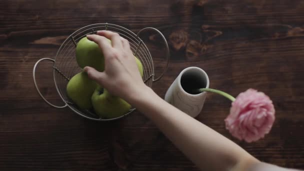 Een jong meisje neemt een appel uit een mand van een tafel en gooit die op een achtergrond van een vaas met een bloem. Een close-up. Uitzicht van bovenaf — Stockvideo