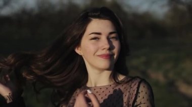 Güzel elbiseli genç bir kız parkta yürüyor ve gülümsüyor. Şık buklelerini elleriyle düzeltiyor. Yakın plan. Yavaş çekim