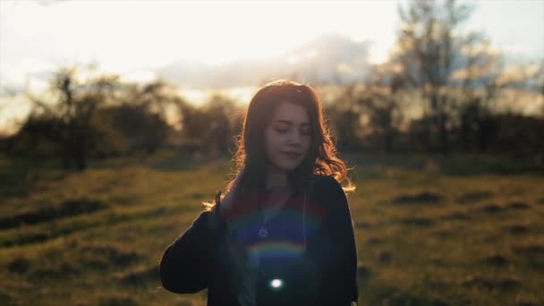 Uma jovem encantadora caminha pelo parque nos raios do pôr-do-sol endireita o cabelo e sorri docemente. Close-up. Movimento lento. Fundo desfocado — Vídeo de Stock