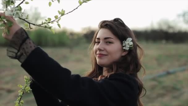 La joven feliz camina en un jardín de primavera floreciente desploma una ramita de flores disfrutando de su aroma e inserta flores en su cabello. Primer plano — Vídeos de Stock