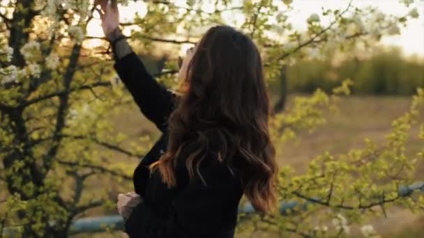 Ein Teenager-Mädchen geht durch einen blühenden Garten in den Strahlen der untergehenden Sonne, schnüffelt Blumen und lächelt glücklich. Nahaufnahme — Stockvideo