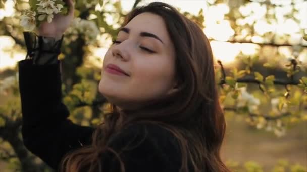 Uma jovem garota atraente em um belo vestido e casaco caminha e gosta do cheiro de jardins florescentes em um dia de primavera. Close-up. Movimento lento — Vídeo de Stock