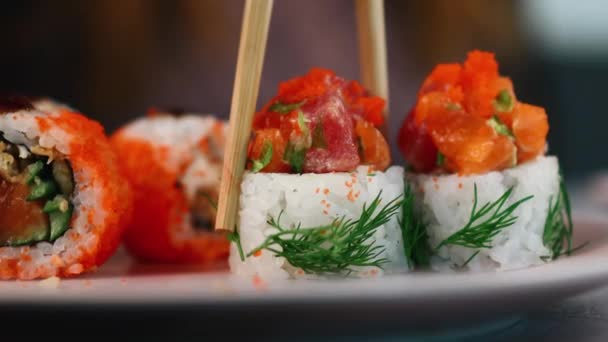 Des rouleaux de sushi japonais orange vif avec du poisson rouge et du caviar et des légumes verts sont pris dans la plaque avec des baguettes. De la nourriture asiatique. Macro shot — Video