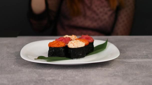 Une jeune fille essaie de prendre de gros sushis Gunkan-maki avec différentes garnitures d'une assiette avec des baguettes. Macro shot — Video