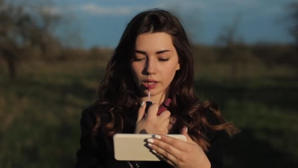 Joven chica risueño hermosa pinta los labios mirando en un teléfono inteligente como si en un espejo caminando en un parque en un día soleado de primavera. Primer plano — Vídeos de Stock