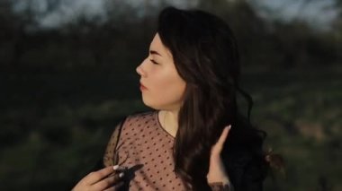 Güzel elbiseli genç bir kız parkta yürüyor ve gülümsüyor. Şık buklelerini elleriyle düzeltiyor. Yakın plan. Yavaş çekim