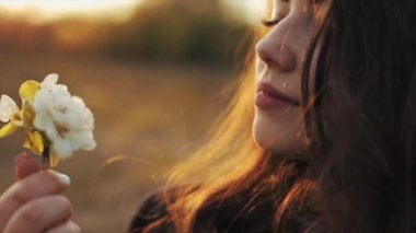 Güzel bir genç kız elinde bir dal tutarak bir elma ağacının çiçeklerini tutar ve batan güneşe karşı bahçede yürürken aromasının tadını çıkarır. Yakın plan. Yavaş çekim