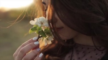 Güzel elbiseli genç bir kız elinde çiçeklerle bir dal tutar ve gözleri kapalı aromayı içine çeker. Yakın plan. Ağır çekim. Kamera odağı değiştirir.