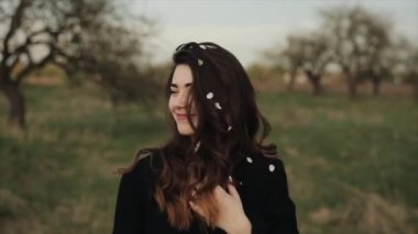 Genç, güzel bir kız çiçek açan bir bahar bahçesinde yürüyüş yapıyor ve saçlarındaki beyaz çiçek yapraklarını silkeliyor. Yakın plan. Ağır çekim. Bulanık arkaplan