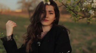 Paltolu güzel bir genç kız bahçede yürürken çiçeklenen dalların arasında duruyor ve saçlarındaki çiçek yapraklarını silkeliyor. Yakın plan.