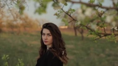 Güzel elbiseli ve ceketli genç çekici kız bahar bahçesinde yürümekten zevk alıyor. Çiçek açan bahçede elleriyle dallara dokunuyor. Yakın plan.