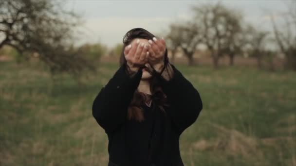 En ung smilende pige i en frakke går på en forårsdag i en blomstrende have og strøede sig selv med blomsterblade. Nærbillede. Langsom bevægelse. Sløret baggrund. Langsom bevægelse – Stock-video