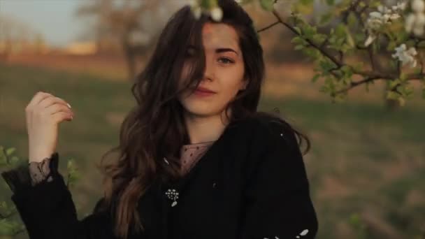 En smuk ung pige i en frakke står blandt blomstrende grene, mens du går i haven og ryster blomsterblade af hendes hår. Nærbillede – Stock-video