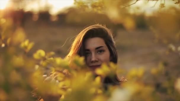 Een jong meisje op een wandeling in de lentetuin staat tussen bloeiende takken en trekt haar haar recht tegen de achtergrond van de ondergaande zon. Een close-up. Langzame beweging. Wazige achtergrond — Stockvideo