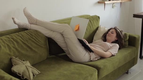 Молодая девушка-фрилансер работает дома на ноутбуке, лежа на диване в своей комнате во время карантина из-за пандемии коронавируса. Крупный план. смешно держит компьютер — стоковое видео
