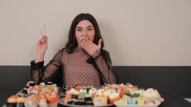 Neşeli, genç, gülümseyen bir kız bir sürü suşinin önünde farklı malzemelerle oturur ve hangisini seçeceğini bilemez. Yakın plan.