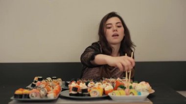 Güzel elbiseli mutlu bir genç kadın, farklı rulo ekmeklerle dolu tabaklarla dolu bir masada sushi yemekten hoşlanır. Yakın plan.