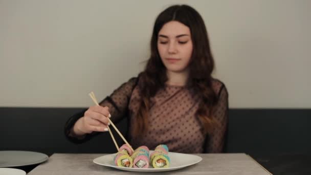 Jong vrolijk aantrekkelijk meisje eet veelkleurige sushi rollen in een café glimlachend en sluit haar ogen van plezier. Close-up — Stockvideo