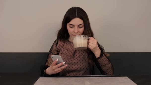 Un'affascinante ragazza beve un caffè in una tazza di vetro seduta a un tavolo in un caffè e chiama al telefono sorridendo. Primo piano — Video Stock