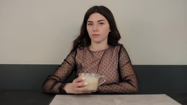 Een jonge glimlachende vrouw drinkt een latte uit een grote glazen beker zittend aan een tafel in een café in een prachtige jurk en kijkt naar de camera. Close-up — Stockvideo